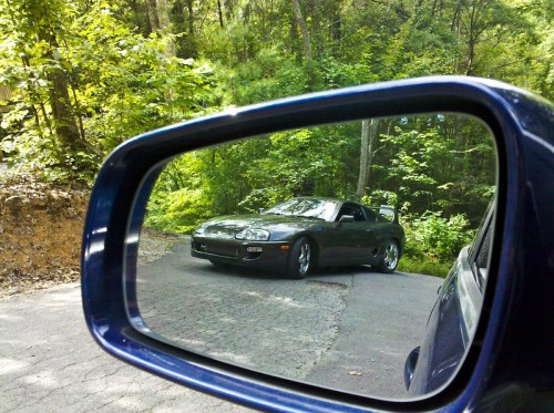 A sleek car parked on a winding road, reflected in the side mirror of another vehicle, surrounded by lush greenery.