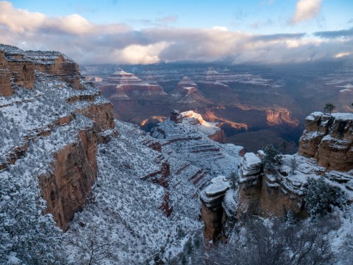 A snowy Grand Canyon landscape with layered rock formations and dramatic clouds in the sky.