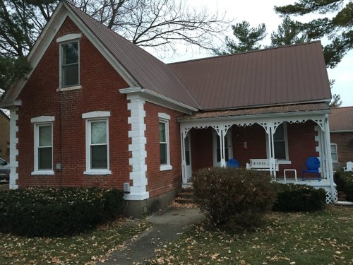 A brick house with a metal roof, featuring a front porch and blue chairs, surrounded by shrubs and fallen leaves.