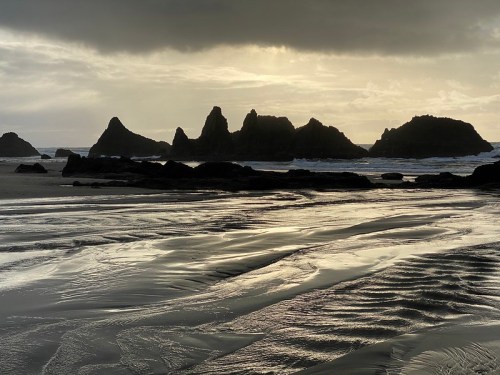Silhouetted rocky formations rise from a sandy beach under a moody, cloudy sky at sunset.