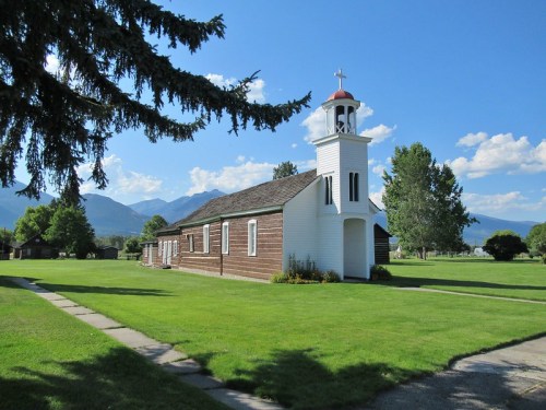 A white church with a red dome, surrounded by green grass and trees, set against a backdrop of mountains and blue sky.