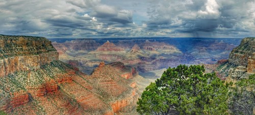A panoramic view of the Grand Canyon, showcasing layered rock formations and dramatic clouds overhead.