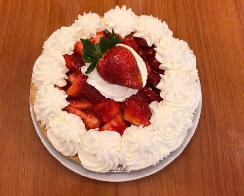A round strawberry cake topped with whipped cream and fresh strawberries, placed on a wooden table.