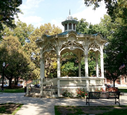A decorative gazebo in a park surrounded by trees and benches, with a clear blue sky above.