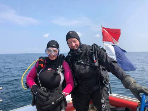 Two divers in wetsuits smile on a boat, with diving flags and the ocean in the background.