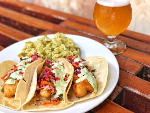 Three tacos with colorful toppings, a side of green rice, and a glass of beer on a wooden table.
