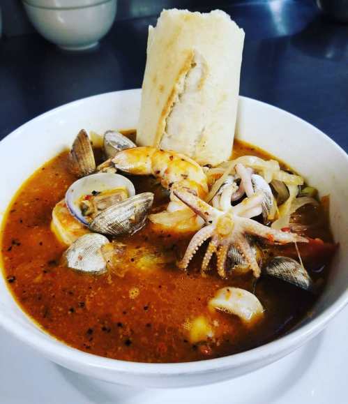 A bowl of seafood soup with shrimp, clams, squid, and a piece of bread, garnished with vegetables in a rich broth.