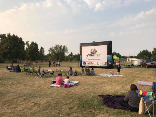 Outdoor movie screening with a large inflatable screen, people sitting on blankets and chairs in a grassy area.