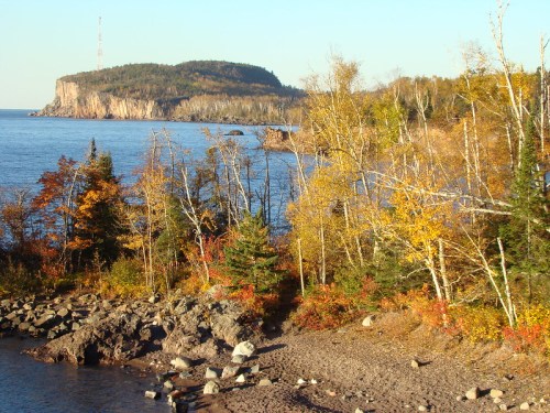 Scenic view of a rocky shoreline with autumn trees and a distant island under a clear blue sky.