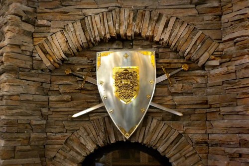 A decorative shield with a lion emblem, flanked by two swords, set against a stone wall.