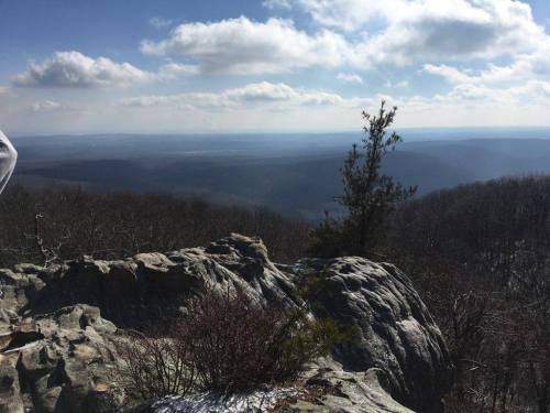 A rocky outcrop overlooks a vast, misty valley with distant mountains under a partly cloudy sky.