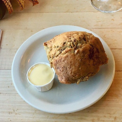 A large, golden-brown muffin on a plate, accompanied by a small cup of creamy sauce.