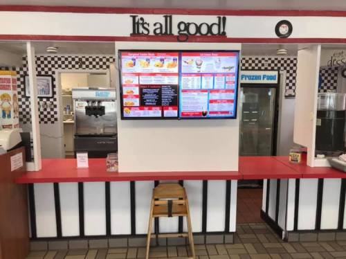A fast food counter with a menu display, red accents, and a sign that says "It's all good!" in a checkered decor.