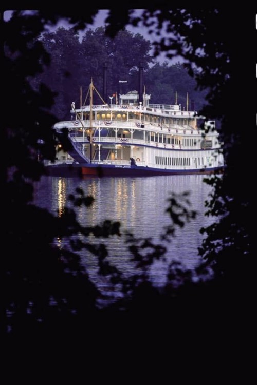 A riverboat illuminated at dusk, framed by trees, reflecting on calm water.