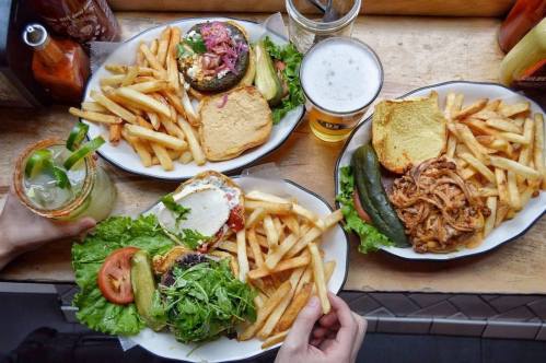 A table with three plates of burgers, fries, and drinks, including a cocktail and beer, in a casual dining setting.