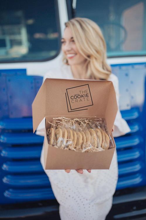 A woman holds an open box of cookies in front of a blue food truck, smiling happily.