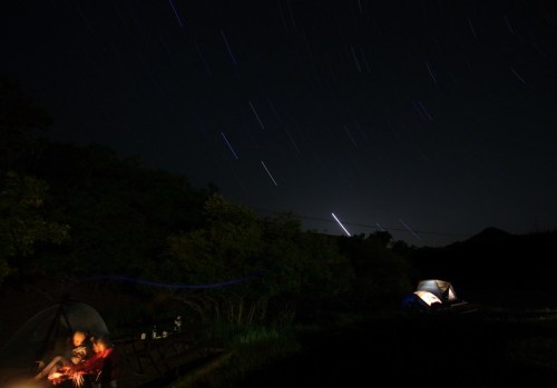 A campsite at night with two tents, a person by a fire, and star trails in the sky.