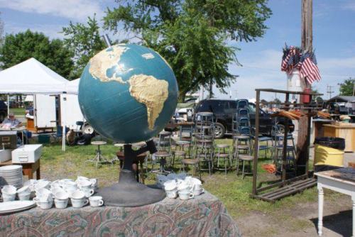 A large globe on a table at a flea market, surrounded by chairs and various items for sale under a clear blue sky.