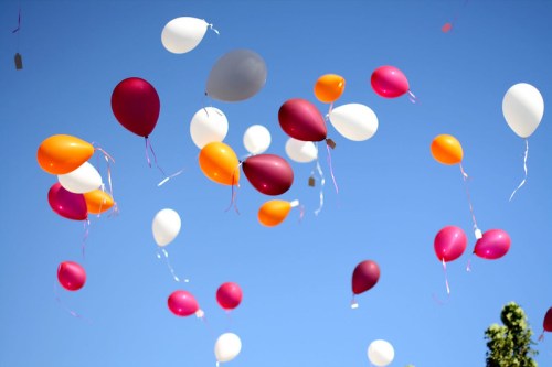 A colorful array of balloons in shades of pink, orange, white, and purple floating against a clear blue sky.