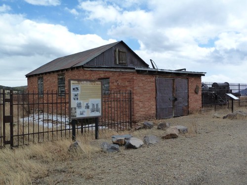A weathered brick building with a metal roof, surrounded by a fence and a sign, set against a cloudy sky.