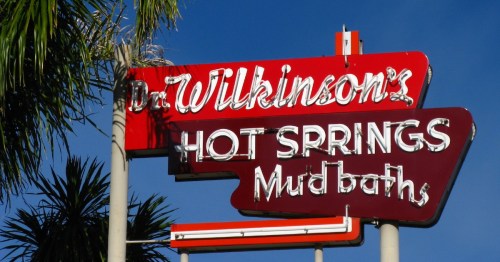 Sign for Dr. Wilkinson's Hot Springs Mud Baths, featuring bold red and white lettering against a blue sky.