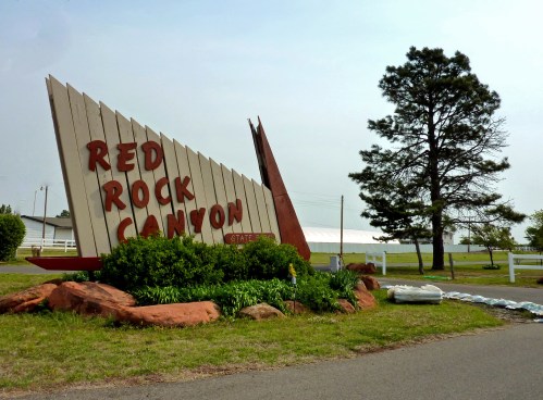 Sign for Red Rock Canyon State Park, featuring bold letters and a natural landscape with greenery and a tree.