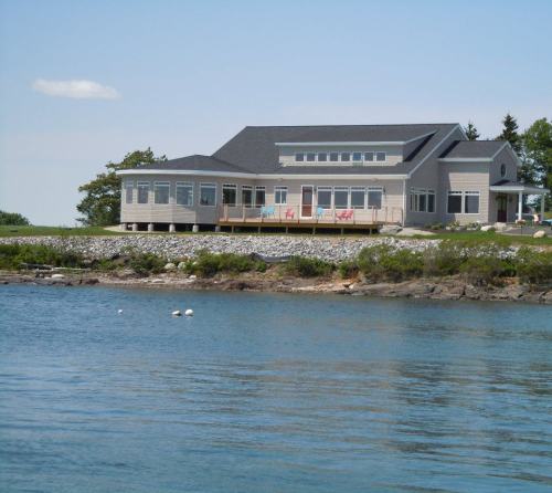 A modern house by the water, featuring a large deck and surrounded by greenery under a clear blue sky.