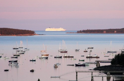A serene harbor at sunset, filled with sailboats, with a large cruise ship in the distance on calm waters.