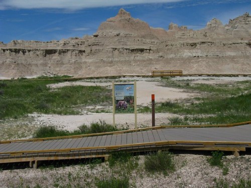 A wooden boardwalk leads through grassy terrain with rocky formations in the background and a sign nearby.