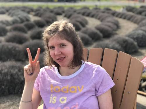 A person sitting in a wooden chair, smiling and making a peace sign, with a field of bushes in the background.