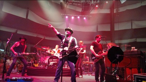 A band performs on stage with colorful lights, featuring a singer with a guitar and musicians playing various instruments.