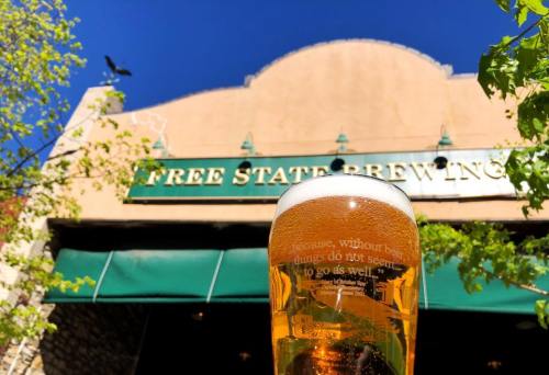 A pint of beer in front of Free State Brewing, with a clear blue sky and greenery in the background.
