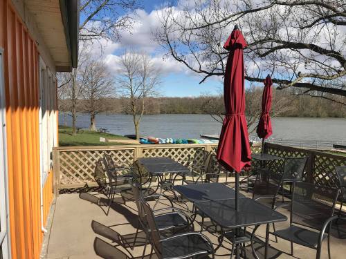 A lakeside patio with black tables and red umbrellas, overlooking a calm water view and trees in the background.