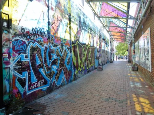 Colorful graffiti covers the walls of a narrow alley, with a brick pathway and a glass-roofed structure above.