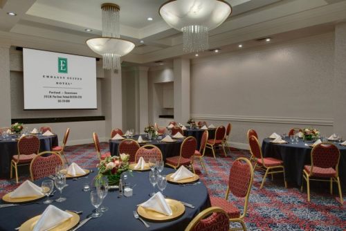 Elegant banquet setup in a hotel conference room with round tables, floral centerpieces, and a projector screen.