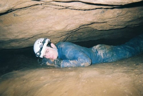 A person in a helmet crawls through a narrow cave passage, covered in dirt and looking towards the camera.