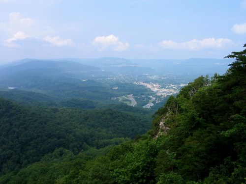 A panoramic view of lush green mountains and a small town nestled in the valley under a blue sky with scattered clouds.