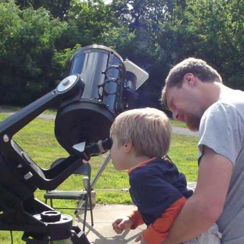A man helps a young boy look through a telescope in a grassy outdoor setting.