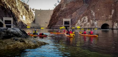 A group of kayakers in orange kayaks paddles near a rocky shore, with a dam visible in the background.