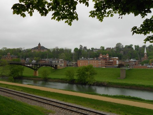 A scenic view of a town with historic buildings, a bridge, and a river, surrounded by green hills under a cloudy sky.
