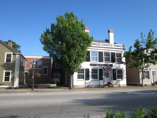 Historic buildings with a large tree in front, decorated with flags, along a quiet street on a sunny day.