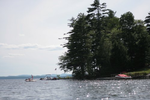 A serene lake scene with a tree-lined shore, boats docked, and a person on a paddleboard in the distance.