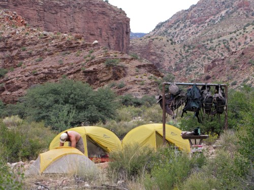 A campsite with two yellow tents in a rocky canyon, a person setting up a tent, and gear hanging on a rack nearby.