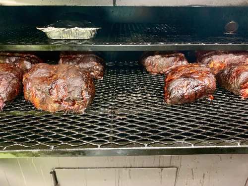 Smoked meats on a grill rack, with a foil tray of food on the top shelf, all showing a dark, charred exterior.