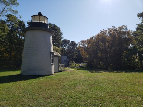 A white lighthouse stands beside a path, surrounded by trees and a clear blue sky on a sunny day.