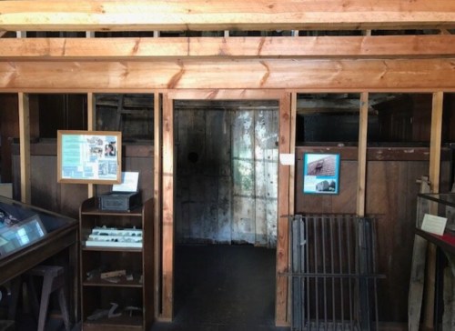 Interior of a wooden building with a doorway leading to a dimly lit room, featuring informational displays on the walls.