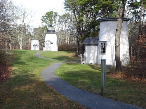 Two white lighthouses on a grassy path, surrounded by trees, with a clear sky in the background.