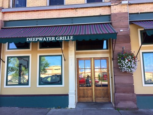 Exterior of Deepwater Grille featuring a striped awning, wooden doors, and flower baskets on a sunny day.