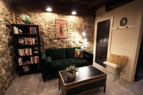 Cozy living room with a green sofa, stone walls, bookshelf, and vintage Houdini poster, featuring warm lighting.