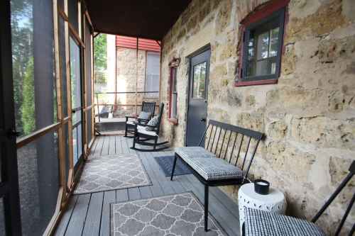 A cozy porch with stone walls, featuring rocking chairs and patterned rugs, surrounded by greenery.
